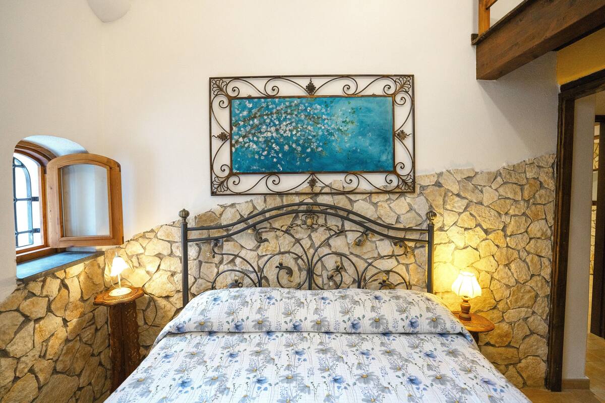 Un dormitorio con pared de piedra, cama con ropa de cama floral, un cuadro sobre la cama y dos lámparas de noche.