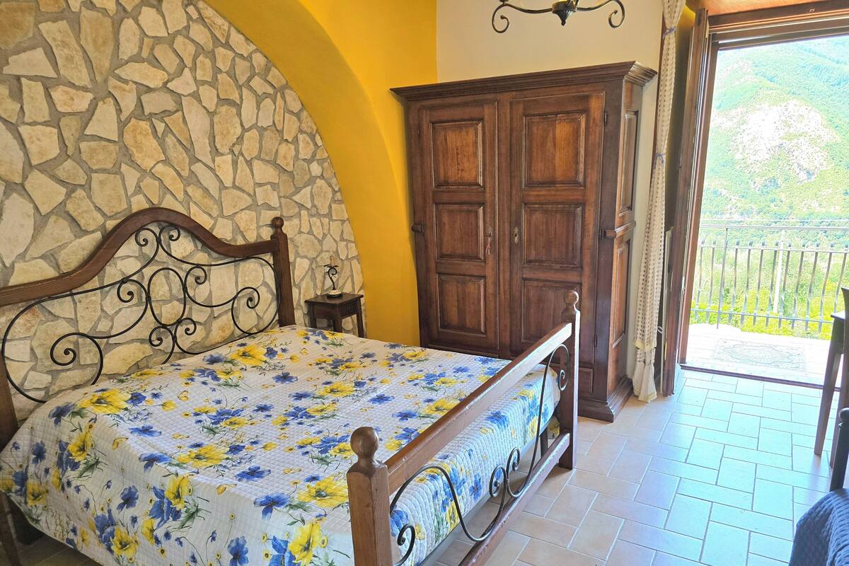 Un dormitorio con pared de piedra, cama de madera con colcha floral, armario de madera y balcón con vistas.