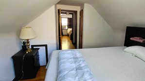 Un dormitorio con cama, mesita de noche con lámpara, cabecero oscuro y vista a un pasillo que conduce a un baño.
