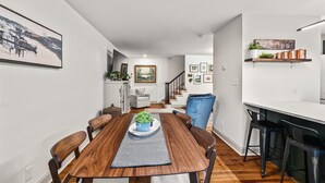 Un comedor moderno con una mesa de madera, sillas y una planta central. Una escalera conduce a una sala de estar con un sofá y una mesa de centro.