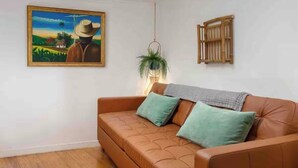 A room with a brown leather sofa, a painting of a rural scene, a hanging plant, and a wooden shutter on the wall.