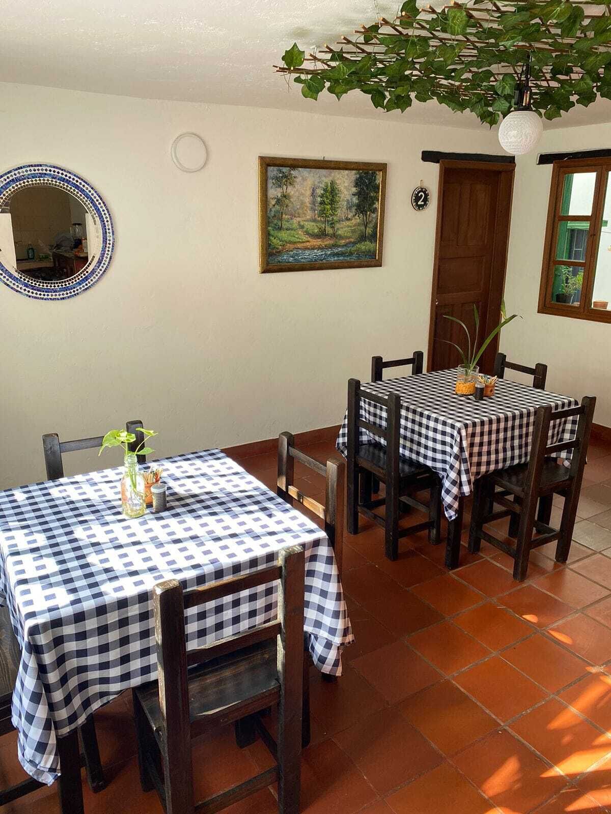A dining area with checkered tablecloths, wooden chairs, a painting on the wall, a mirror, and a hanging plant.