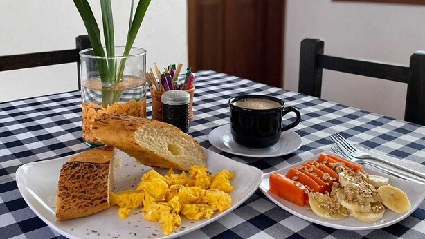 Una mesa de desayuno con mantel a cuadros, platos de huevos revueltos, tostadas, fruta, una taza de café y un florero con plantas verdes.