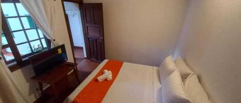 A single bed with white linens and an orange accent, a wooden bench, a window with curtains, and a door in a room with plain walls.