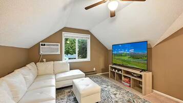 Un salon avec un canapé d’angle blanc, un petit pouf, un téléviseur à écran plat posé sur un meuble en bois, un ventilateur de plafond et une fenêtre donnant sur un paysage verdoyant.
