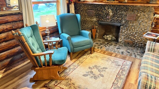 L’intérieur d’un chalet confortable, avec une cheminée en pierre, un fauteuil bleu, une chaise à bascule en bois et un canapé à carreaux.