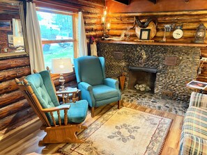 Ein gemütliches Blockhaus-Interieur mit Kamin aus Stein, einem blauen Sessel, einem hölzernen Schaukelstuhl und einem karierten Sofa.