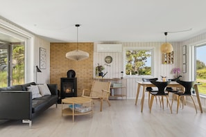 A modern living room with a fireplace, a dining area with a table and chairs, and large windows offering a view of greenery.