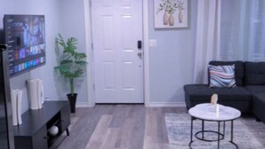 A modern living room with a grey floor, a white door, a black sofa, a coffee table, and a TV on a stand. - Cozy 3 BR Family Home Near OU Hospital (Oklahoma City)