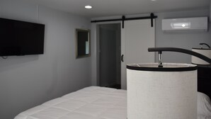Una habitación de hotel con cama, televisor montado, aire acondicionado instalado en la pared y lámpara de noche.