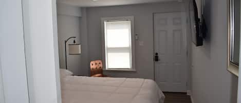 A bedroom with a bed, a chair, a window, a door, and a mounted TV.