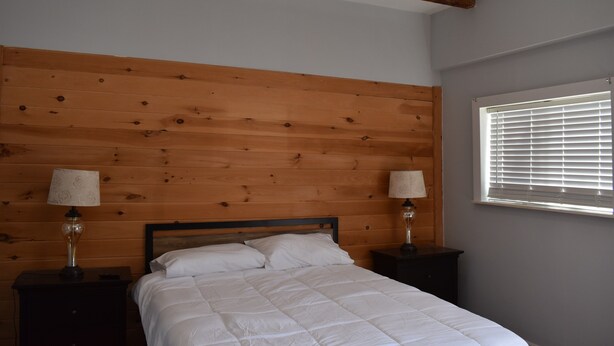 Une chambre à coucher avec une tête de lit en bois, un lit avec une literie blanche, deux tables de chevet avec des lampes et une fenêtre avec des stores.