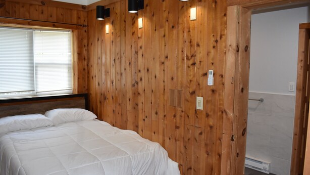 A bedroom with a wooden wall, a bed with white bedding, a window with blinds, and a door leading to a bathroom.
