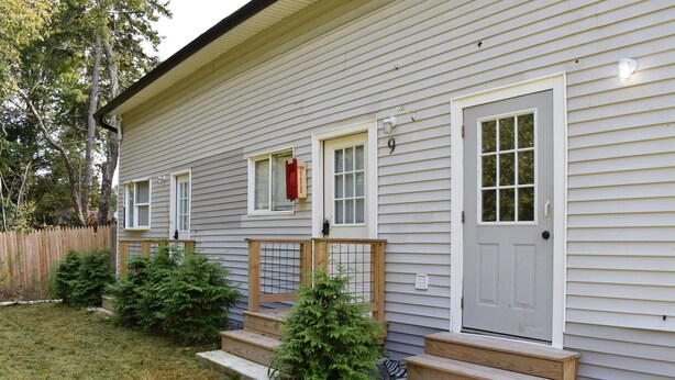 Une maison à deux étages, avec une façade grise, des éléments décoratifs blancs et le chiffre « 9 » sur la porte. Des marches mènent à la porte d’entrée, et un petit arbre est planté devant.