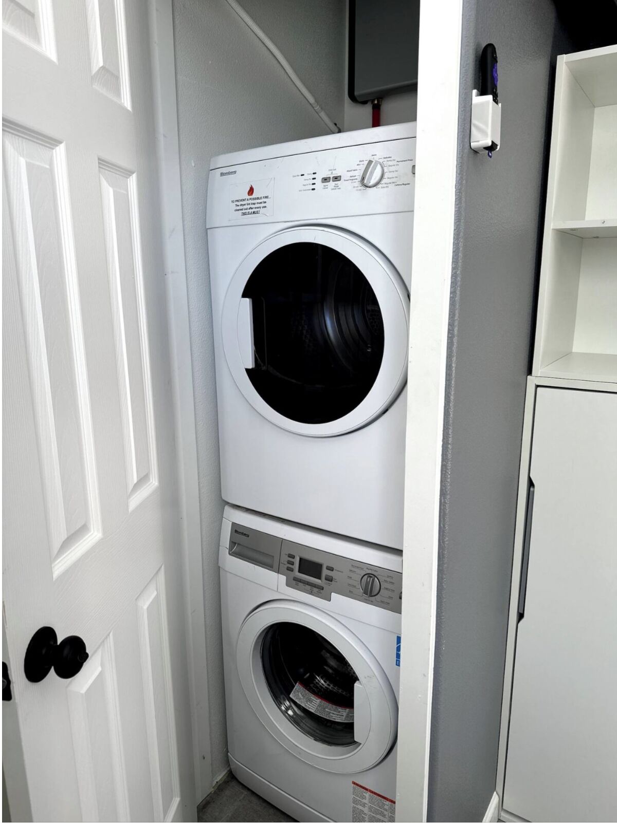 Une petite buanderie équipée d’un lave-linge et d’un sèche-linge superposés, d’une porte blanche et d’une petite étagère.