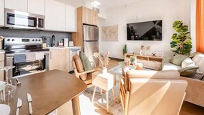 A modern kitchen with stainless steel appliances, a dining area with a wooden table and chairs, and a living room with a sofa and a TV.