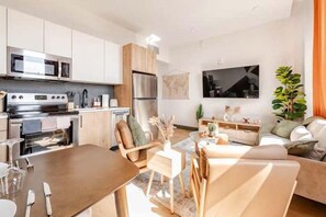 A modern kitchen with stainless steel appliances, a dining area with a wooden table and chairs, and a living room with a sofa and a TV.