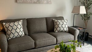 A grey sofa with patterned cushions, a wall-mounted artwork of branches, a floor lamp, and a small table with a magazine.