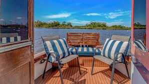 Ein Bootsdeck mit einer Holzbank und gestreiften Kissen, Blick auf Wasser und Grünflächen.
