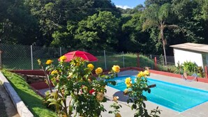 A swimming pool surrounded by a fence, with a red umbrella and yellow flowers in the foreground. - Recanto Raios do Sol  (Caldas)