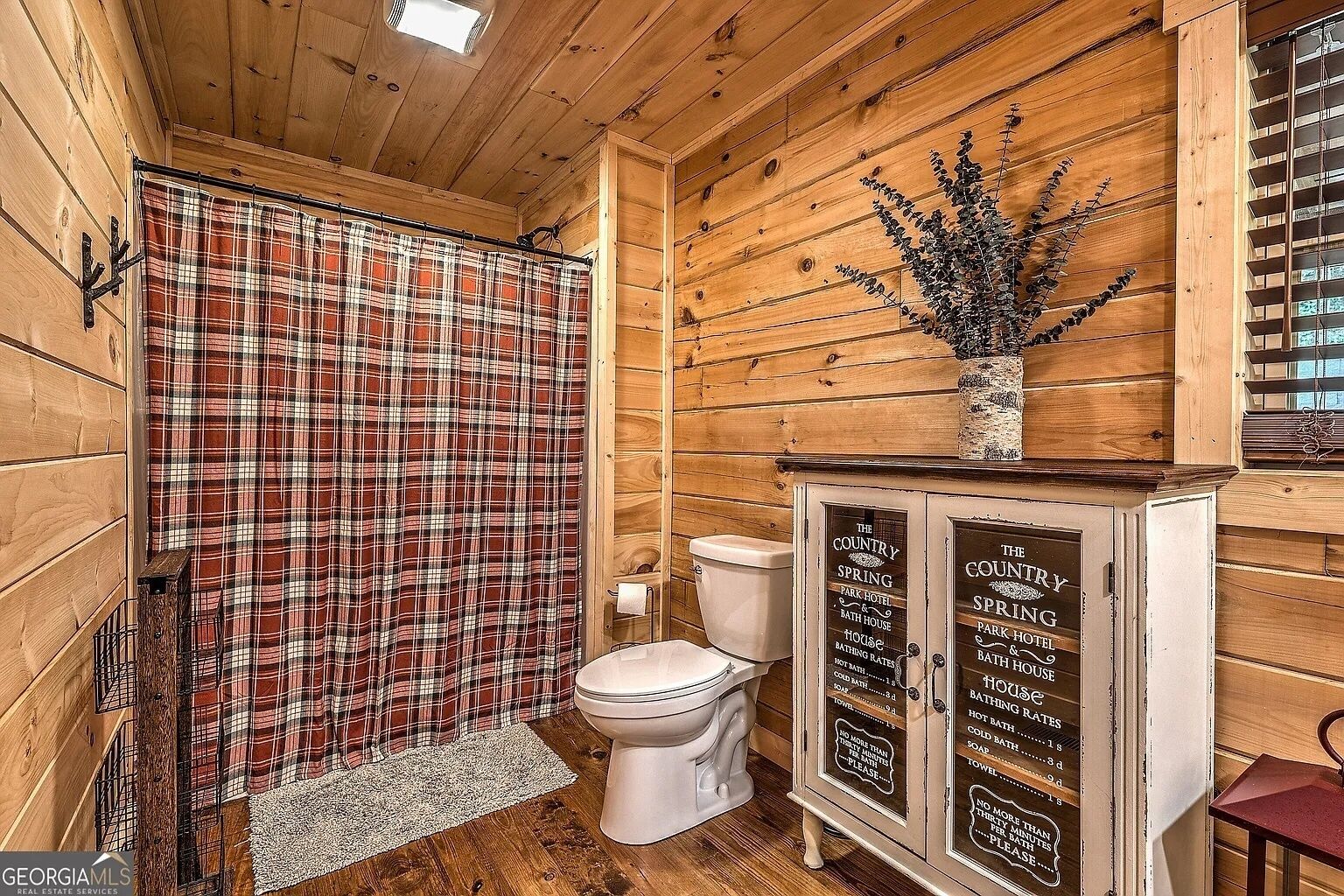Een rustieke badkamer met een geruit douchegordijn, een toilet en houten kastjes met een bord waarop faciliteiten staan vermeld, zoals een parkhotel en badhuis.