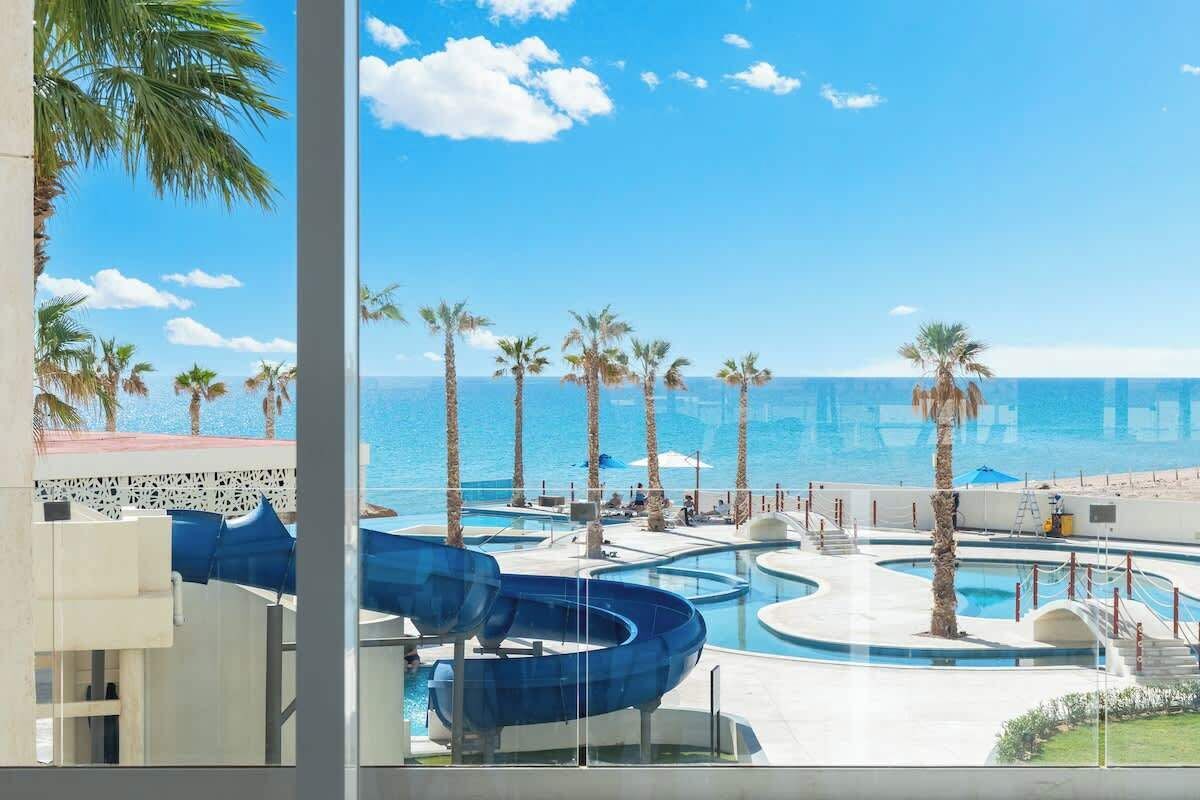 Ein Poolbereich mit mehreren Wasserrutschen und Palmen, mit Blick auf einen Strand.
