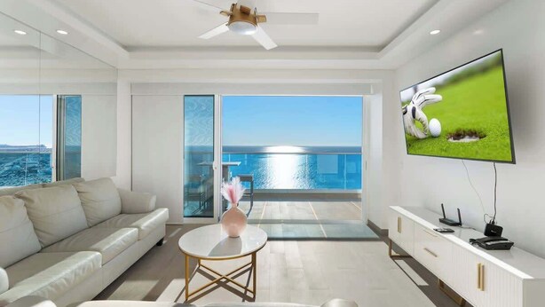 A modern living room with a sofa, a small table, a TV, and a balcony with a view of the sea.