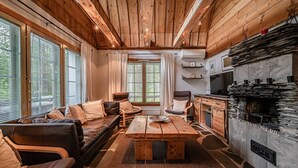 A cozy cabin interior with a wooden ceiling, a stone fireplace, a sofa, a coffee table, and a kitchen area.