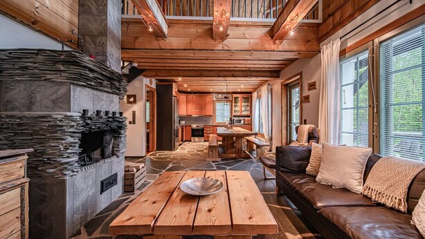 A spacious living area with a stone fireplace, wooden ceiling beams, and a large window providing natural light.