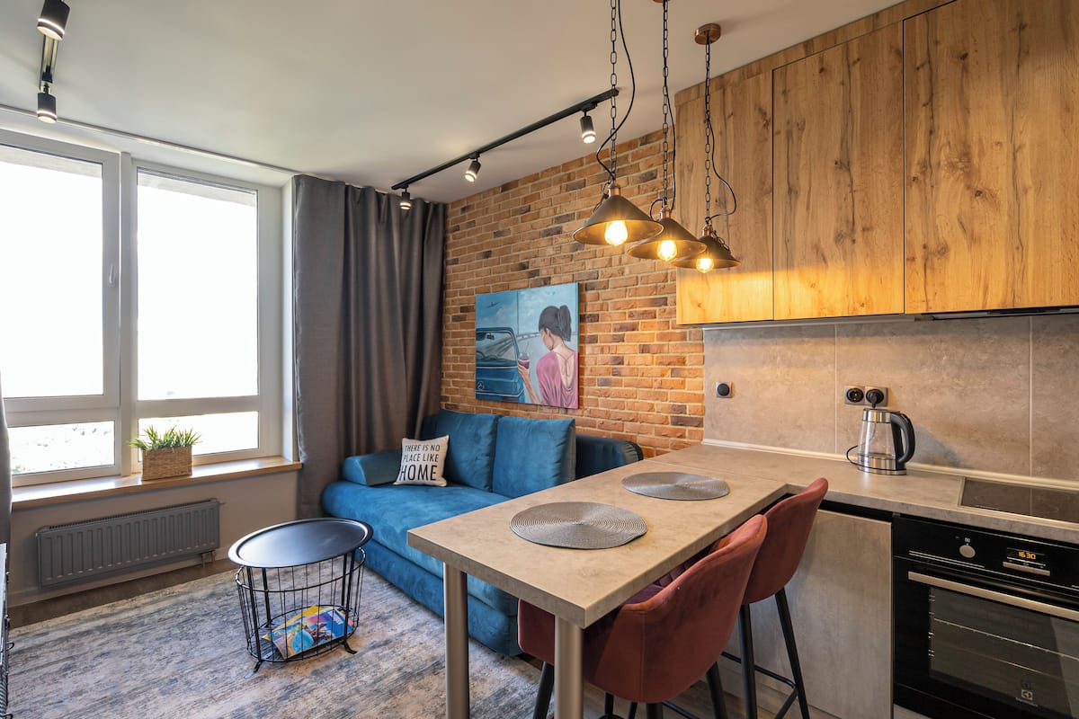 A modern kitchen with wooden cabinets, a dining area with a table and chairs, a blue sofa, and a radiator near the window.