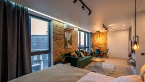 Une chambre d’hôtel moderne dotée d’une grande fenêtre, d’un mur décoratif en briques, d’un canapé vert, d’une petite table ronde et d’un lit avec une literie blanche.