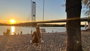 Une plage avec une balançoire et un arbre, un pont en arrière-plan et le soleil se couchant sur l’eau.