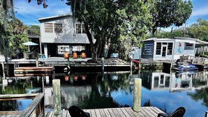 Un muelle de madera con sillas, una casa y un barco atracado junto a una masa de agua tranquila.