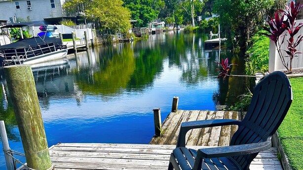 Un muelle de madera con dos sillas Adirondack mirando un cuerpo de agua tranquilo, rodeado de follaje y una casa al fondo.