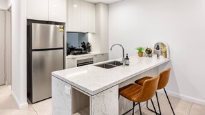 Une cuisine moderne avec des meubles blancs, un réfrigérateur en acier inoxydable, un comptoir en marbre et deux tabourets de bar marron.