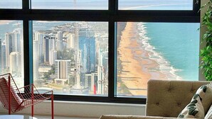 A room with a large window overlooking a beach and cityscape, a red chair, and a beige sofa.