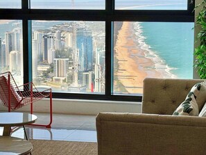Ein Zimmer mit großem Fenster, das Blick auf Strand und Stadt bietet, ein roter Sessel und ein beiges Sofa.