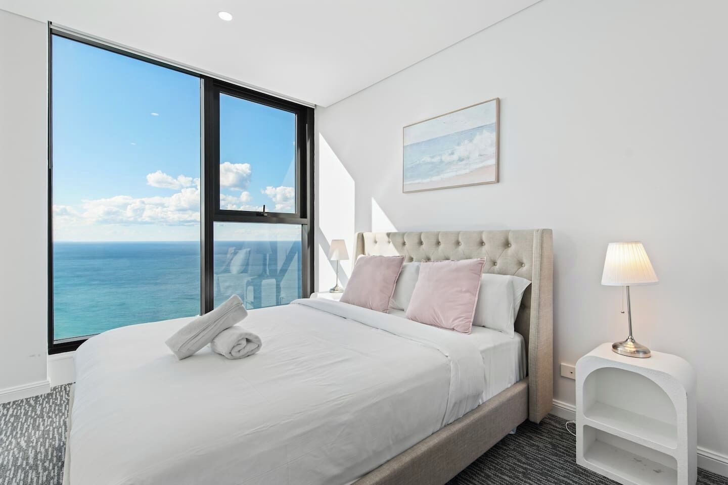 A bedroom with a large window, a bed with white bedding, a tufted headboard, a bedside lamp, and a framed picture on the wall.