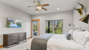 Un dormitorio con cama, ventilador de techo, televisor montado, ventana con vistas y lámpara de noche.