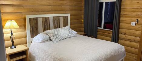 A wooden cabin bedroom with a bed, a nightstand, a lamp, and a window with curtains.