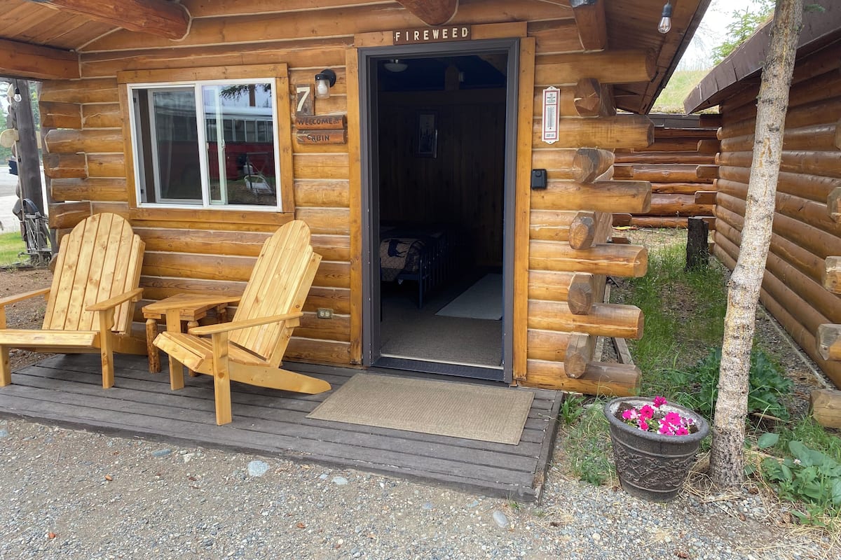 A log cabin with a wooden deck, two Adirondack chairs, a welcome sign, and a cabin number sign.