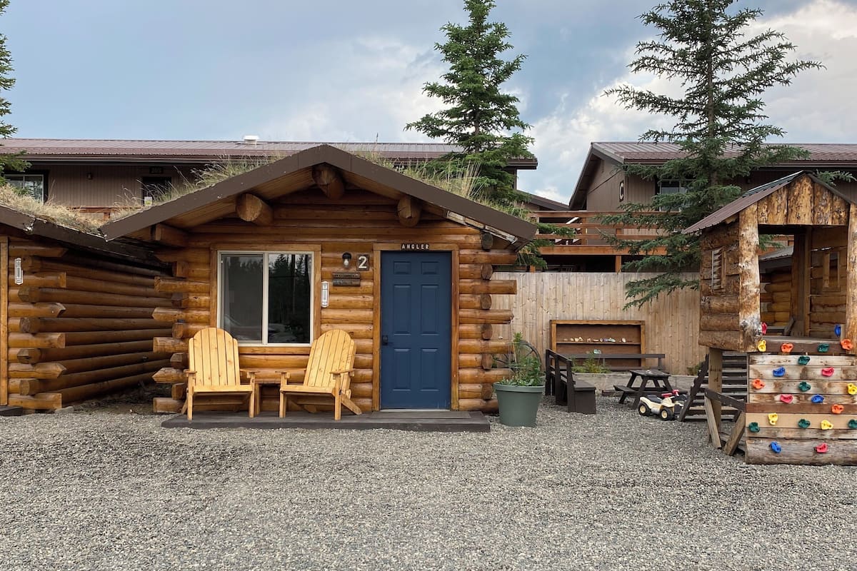 A rustic log cabin with a blue door, numbered 2, and two wooden Adirondack chairs in front.