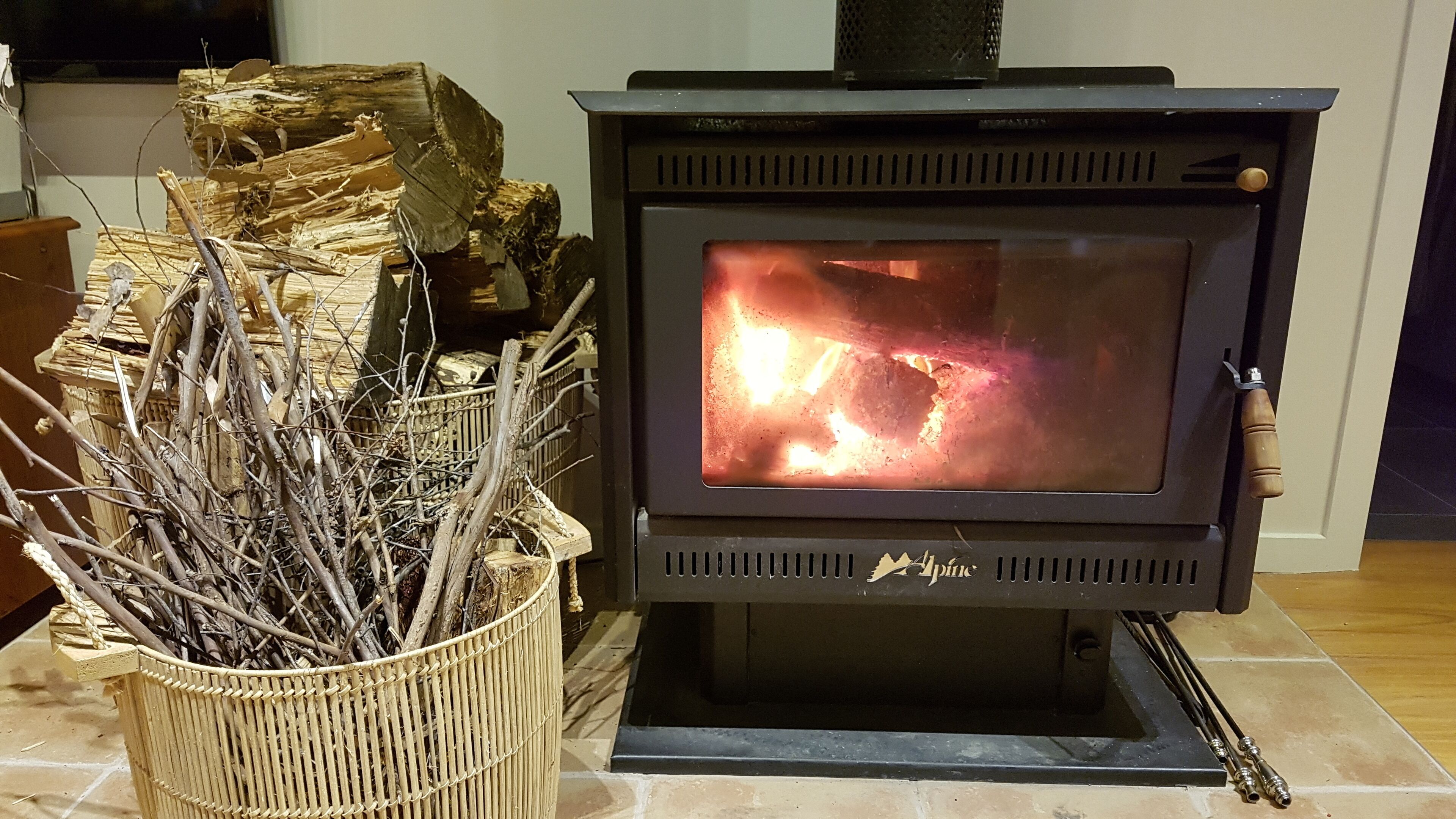 A wood-burning stove with visible flames, a wicker basket with branches, and a stack of firewood.
