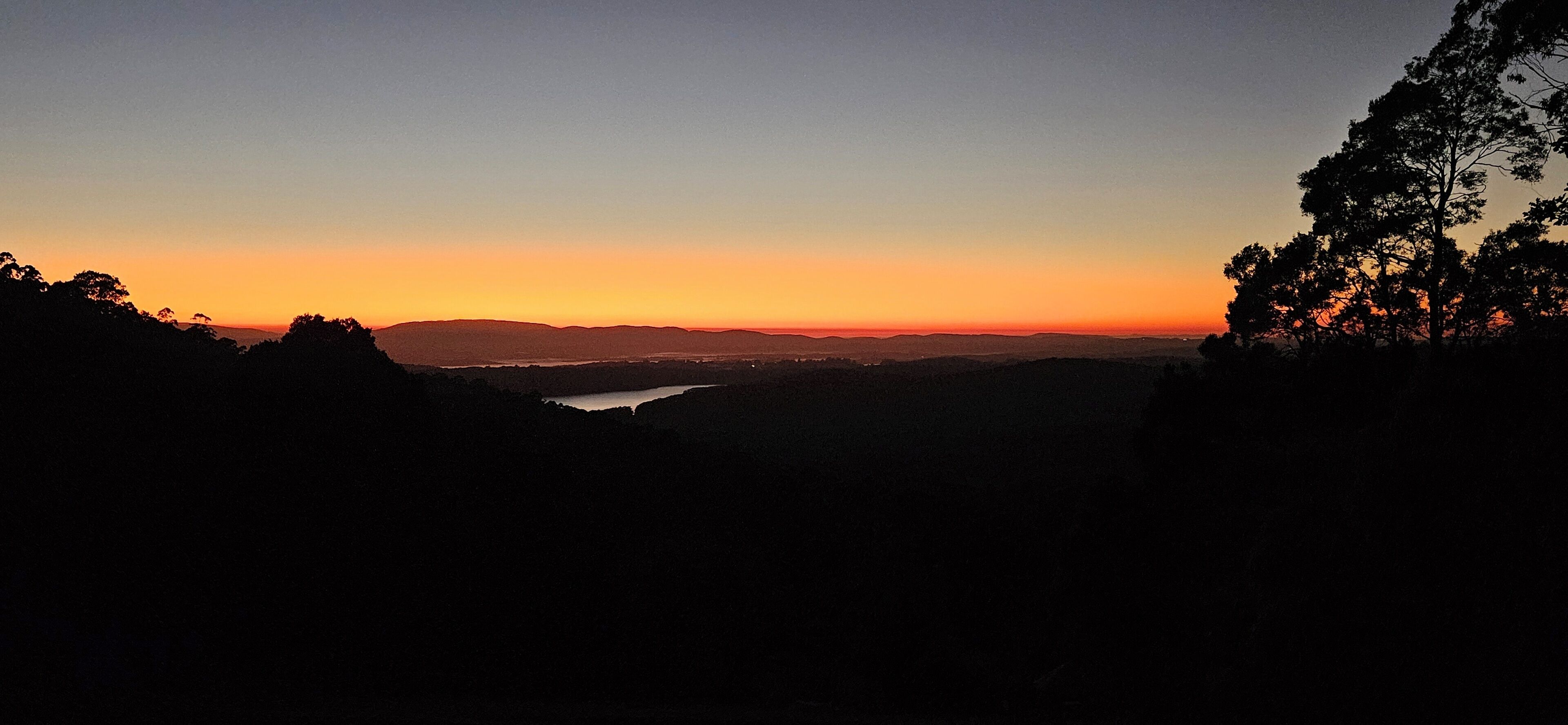 Vista al atardecer desde un punto elevado con vistas a un valle, colinas lejanas y una masa de agua.