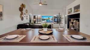 A dining area with a long table set for four, a ceiling fan, and a TV displaying a tropical scene. - Azure Oasis - Saltwater Pool/Spa with putting green! (Palm Desert)