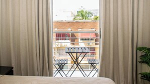 Een slaapkamer met een groot raam, gordijnen, een bed en uitzicht op gebouwen en een balkon.
