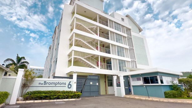 Een modern meerlaags gebouw met een opvallende ingang, een bord waarop “the Bromptons 6” staat en een hekwerk oprit.