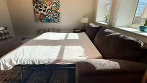 A bedroom with a bed, a brown sofa, a window with a view, and a colorful abstract painting on the wall.
