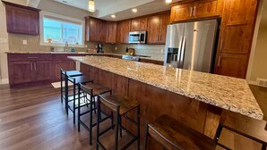 A modern kitchen with wooden cabinets, a granite countertop, stainless steel appliances, and bar stools.