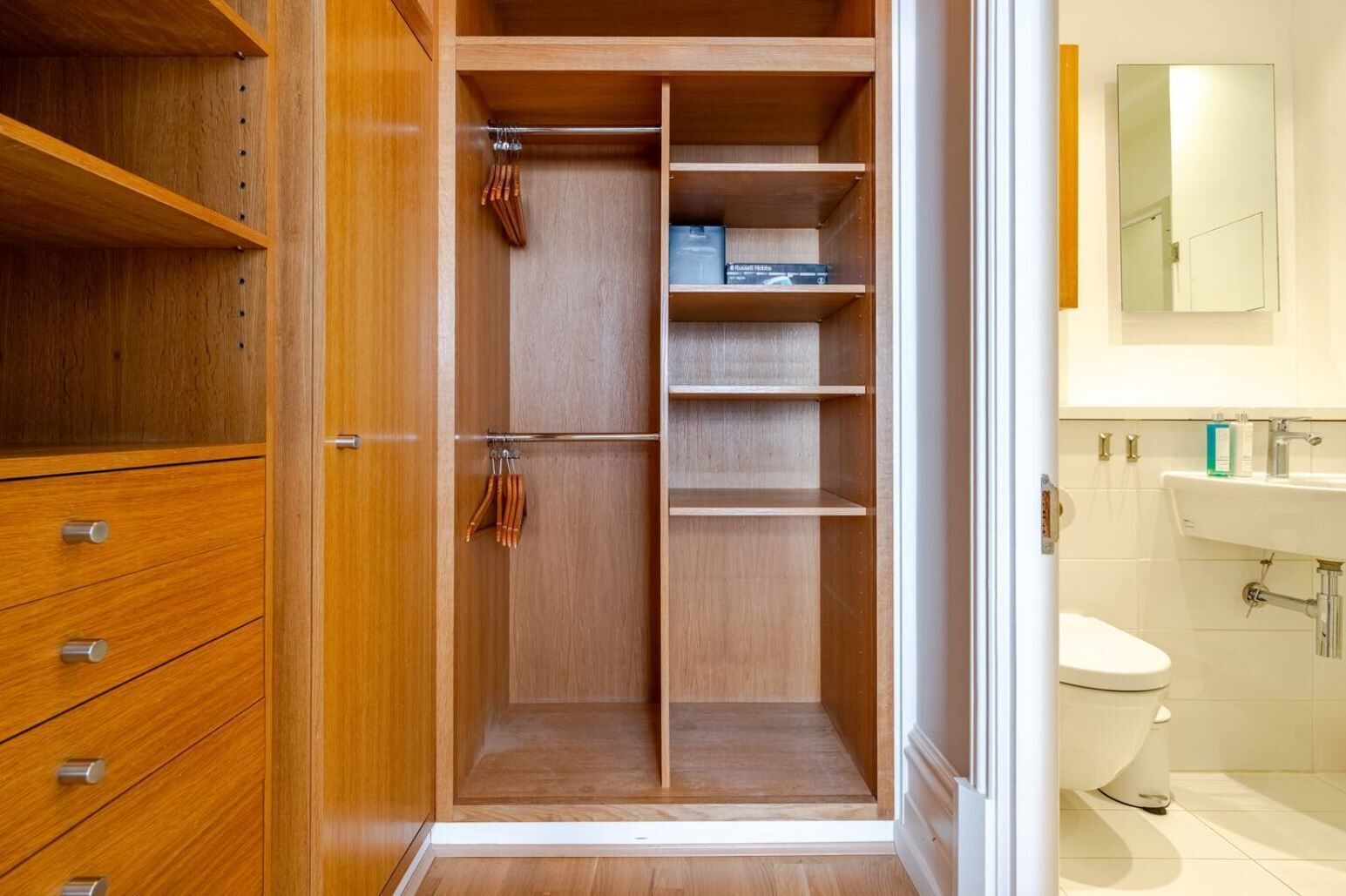 A walk-in closet with wooden shelving, a hanging rod, and a door leading to a bathroom with a toilet and sink.